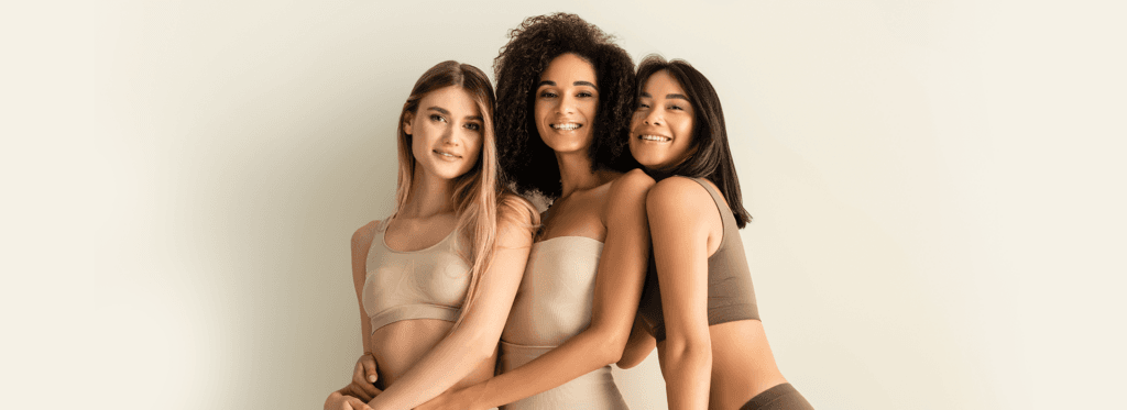 Três mulheres diversas sorrindo e se abraçando, mostrando a beleza natural e positividade corporal.
